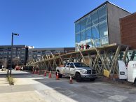 Construction workers stand on a wooden scaffolding outside a modern glass building. Orange cones and a white pickup truck block off the sidewalk and part of the street. An apartment building appears in the background.