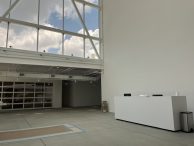 A modern, spacious lobby with high ceilings and large windows showing a partly cloudy sky. There is a white reception desk, a trash bin, and a cardboard box near the wall on the right.