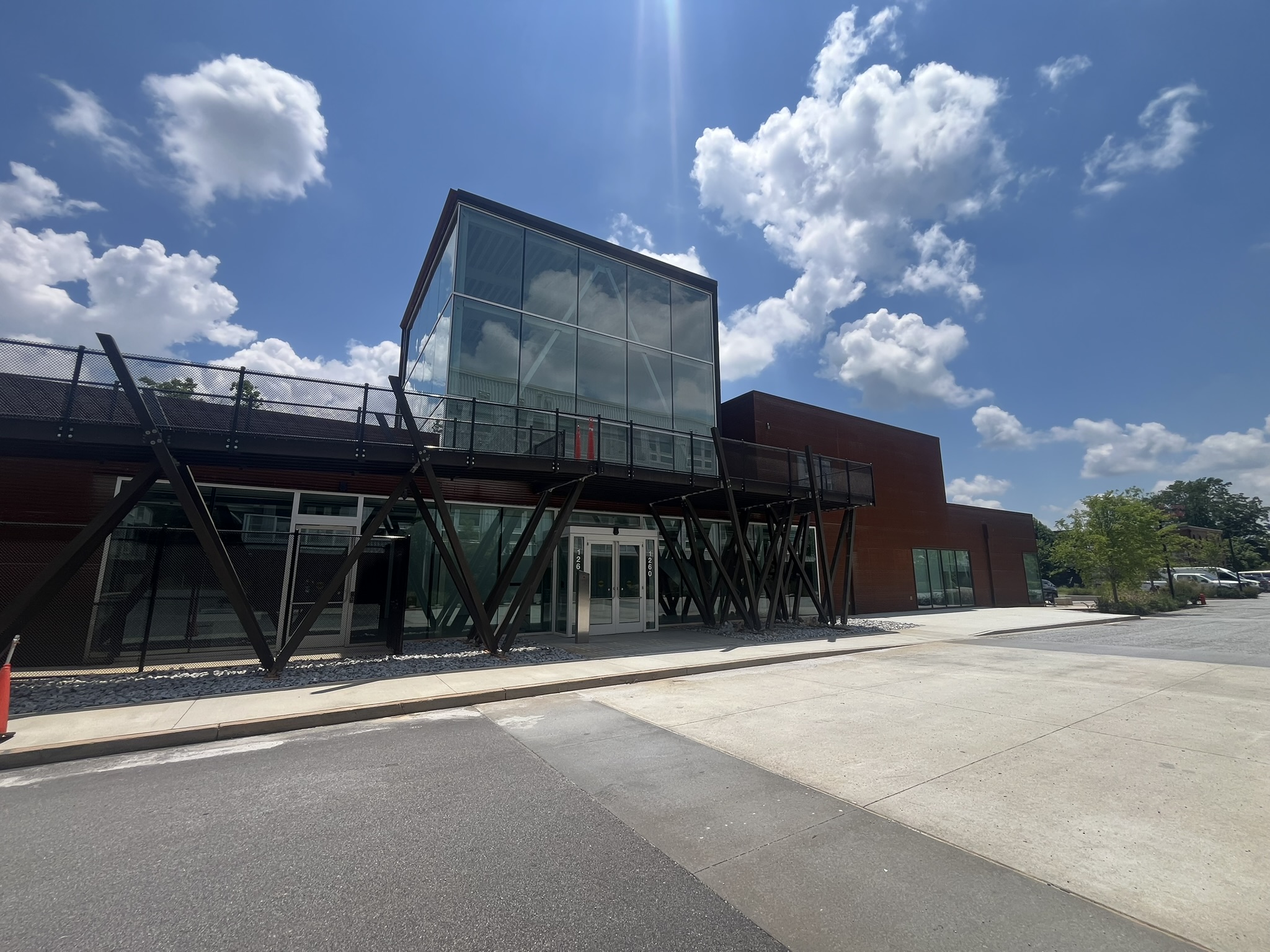 Modern building with large glass windows and unique black metal supports in a V-shape, set against a bright blue sky with scattered clouds. The sidewalk and street are visible in the foreground.