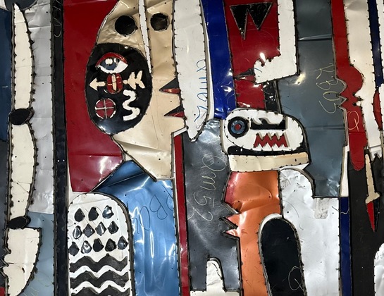 Colorful abstract metal art features jagged, face-like shapes with bold lines, geometric patterns, and symbols. The artwork has a textured, layered appearance, with reds, blues, and whites dominating the scene.