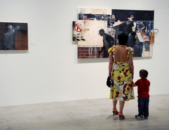 A woman in a yellow floral dress and a child in a red shirt stand in an art gallery, looking at modern paintings displayed on a white wall.