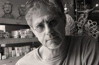 A man with glasses and light hair stands in a workshop surrounded by shelves filled with art supplies and sculptures. The image is in black and white.