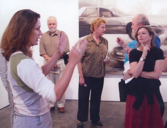 A woman gestures while speaking to a group of attentive adults in an art gallery. A blurred painting of a car is visible on the wall behind them.