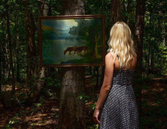 A woman with blonde hair in a patterned dress stands in a sunlit forest, looking at a framed painting of horses by a lake that is hanging on a tree.
