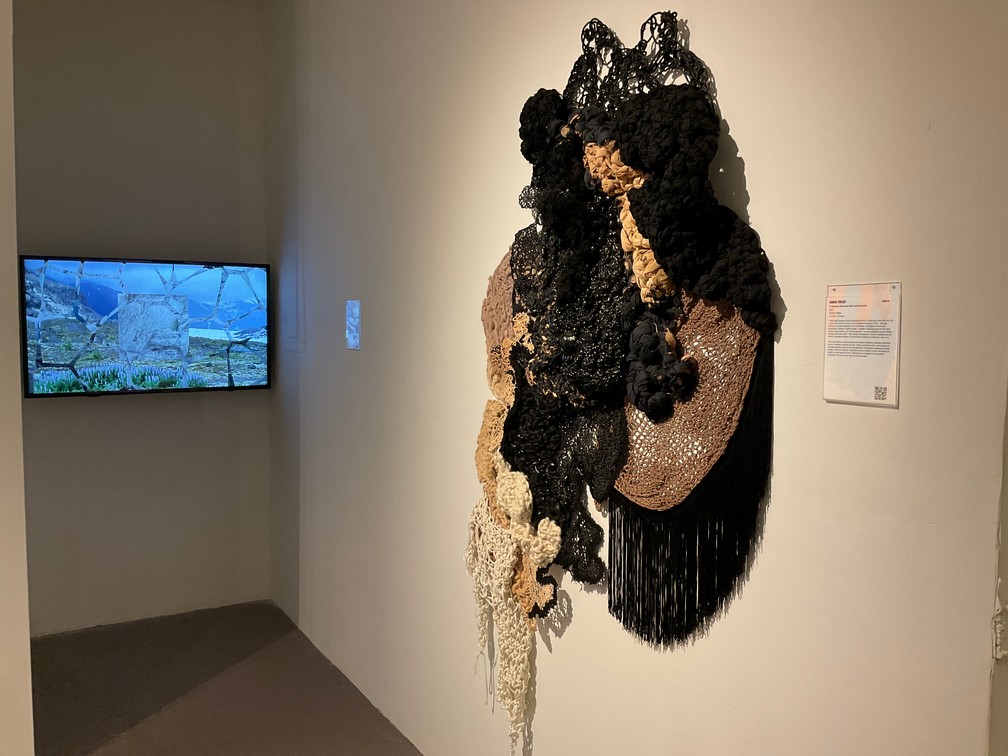 Textured black, beige, and white wall sculpture hangs in a gallery next to an information plaque. In the background, a digital screen displays a landscape image on the adjacent wall.
