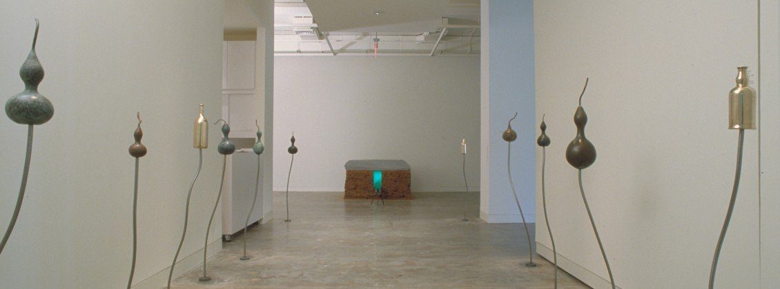A minimalist gallery with curved metal stems holding gourd-shaped sculptures lines both sides of a polished hallway, leading to a rectangular bench with a glowing green light underneath.