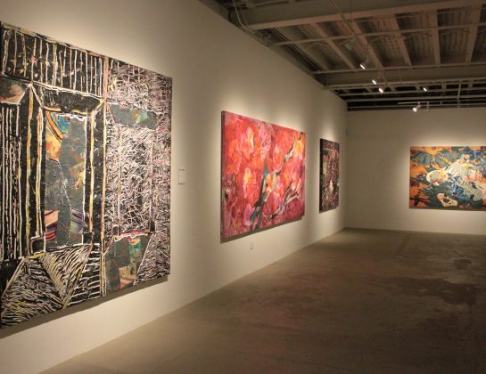 A contemporary art gallery with three large colorful paintings displayed on white walls, featuring abstract and floral designs under ceiling spotlights. Concrete floors and a high, open ceiling create a modern atmosphere.