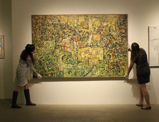 Two people wearing gloves adjust a large, colorful abstract painting on a gallery wall. The painting features swirling patterns and intricate details.