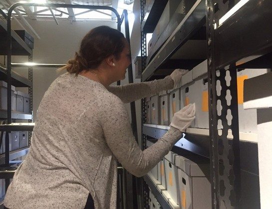 A person wearing white gloves reaches for a storage box on a tall metal shelving unit.