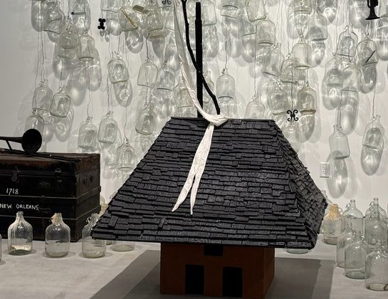 An art installation featuring glass jugs, a slate-roofed model house with fabric tied on top, a black chest, a small black sailboat on a red table, and more glass jugs arranged on the floor and hanging in the background.